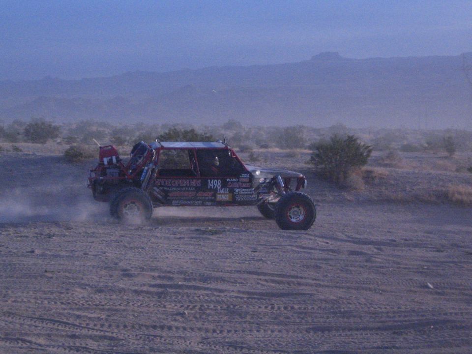 Off-road race buggy number 1498 throwing dust on desert flat at dusk
