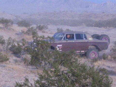 Maroon vintage luxury sedan converted to off-road desert racer parked on brush-covered hillside