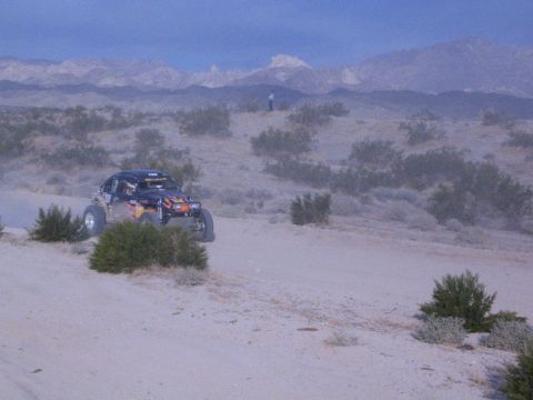 Dark off-road buggy on sandy desert trail with snow-dusted mountains and a spectator
