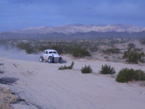 White off-road racing car raising dust on desert berm with snow-dusted mountain range