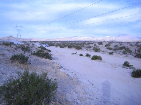 Distant off-road car raising dust on desert flats beneath power lines and mountains