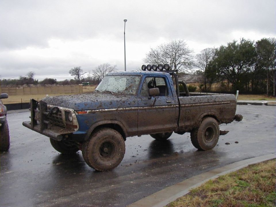 Mud-covered blue pickup truck with a bull bar and roof lights parked on a wet road
