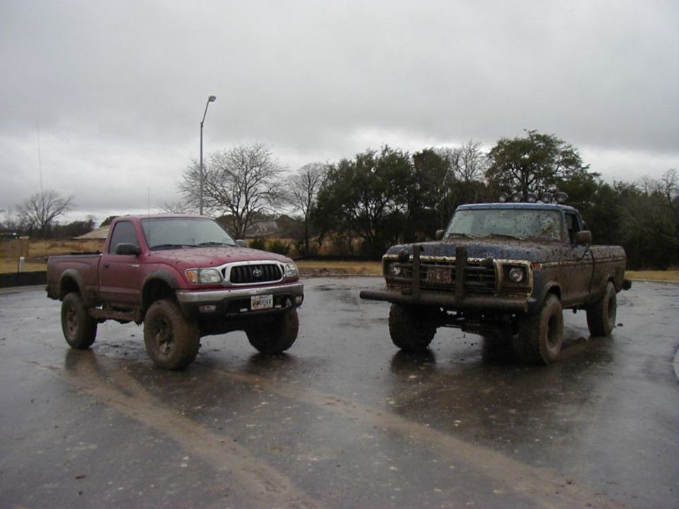 Two muddy pickup trucks, one red Toyota Tacoma and one dark blue older model Ford pickup, parked on wet pavement