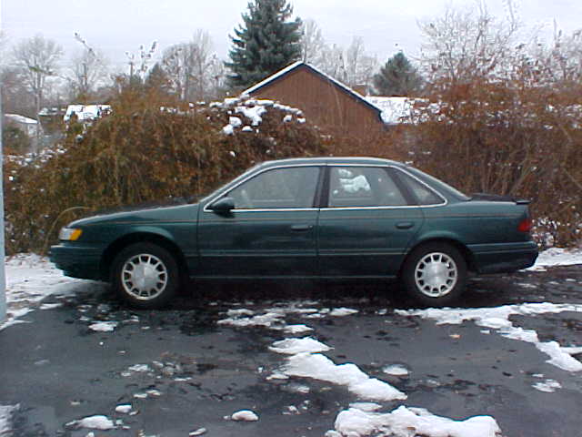 Green Ford Taurus sedan parked sideways on driveway before hedge, light snow on ground
