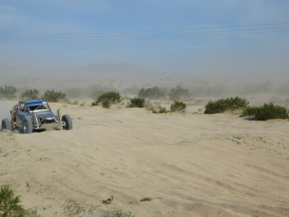 Blue off-road buggy cresting a sandy rise in a dusty desert landscape