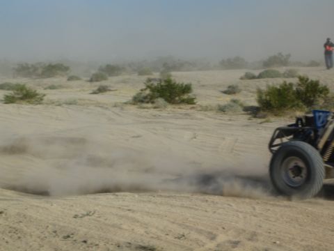 Rear of a dark off-road buggy raising dust on sand with a flagger standing in the background