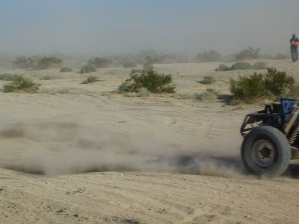 Rear of a dark off-road buggy raising dust on sand with a flagger standing in the background