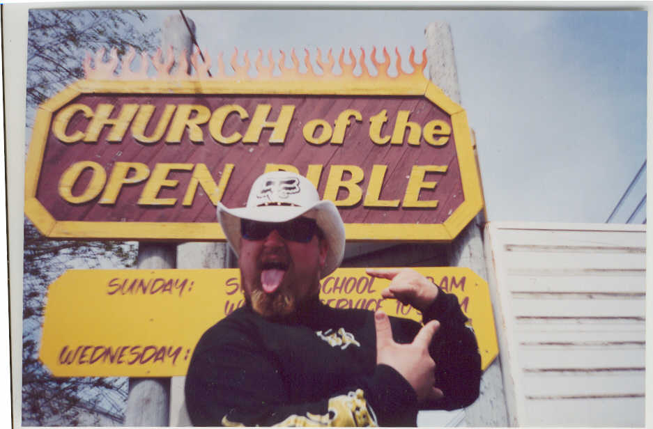 Man in cowboy hat and sunglasses posing under a flame-trimmed Church of the Open Bible sign