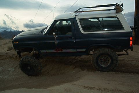 1986 Ford Bronco - This 1986 Ford Bronco is shown off-roading in a sandy, mountainous terrain under a cloudy sky.