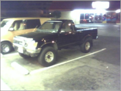 1989 Mazda MX-6 - A black pickup truck is parked in a lot next to a tan van, with a building in the background.