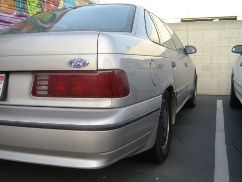 1991 Ford Taurus - The rear of a silver sedan is shown, parked in a lot next to another vehicle.