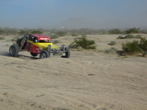 Magenta and yellow desert race buggy racing across sand under a blue sky