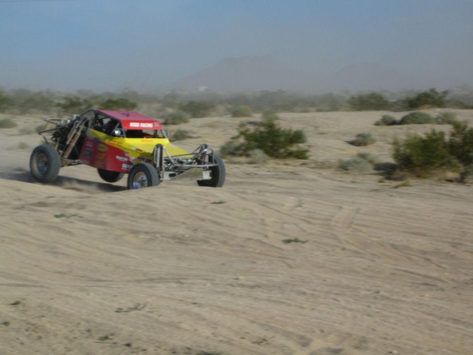 Magenta and yellow desert race buggy racing across sand under a blue sky
