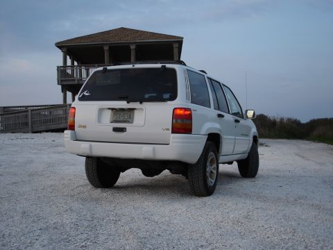1997 Jeep Grand Cherokee - A white 1997 Jeep Grand Cherokee is viewed from the rear on a gravel lot with a beach house in the background.