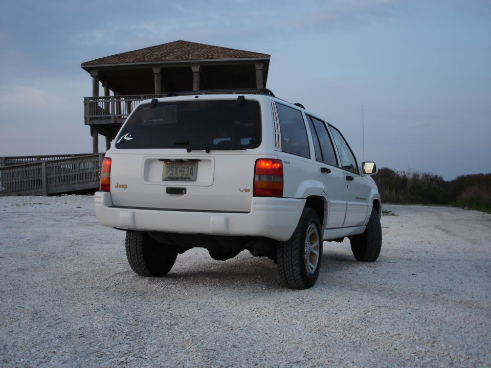 White four-door Jeep SUV with V8 badge parked on a shell lot near a beach pavilion