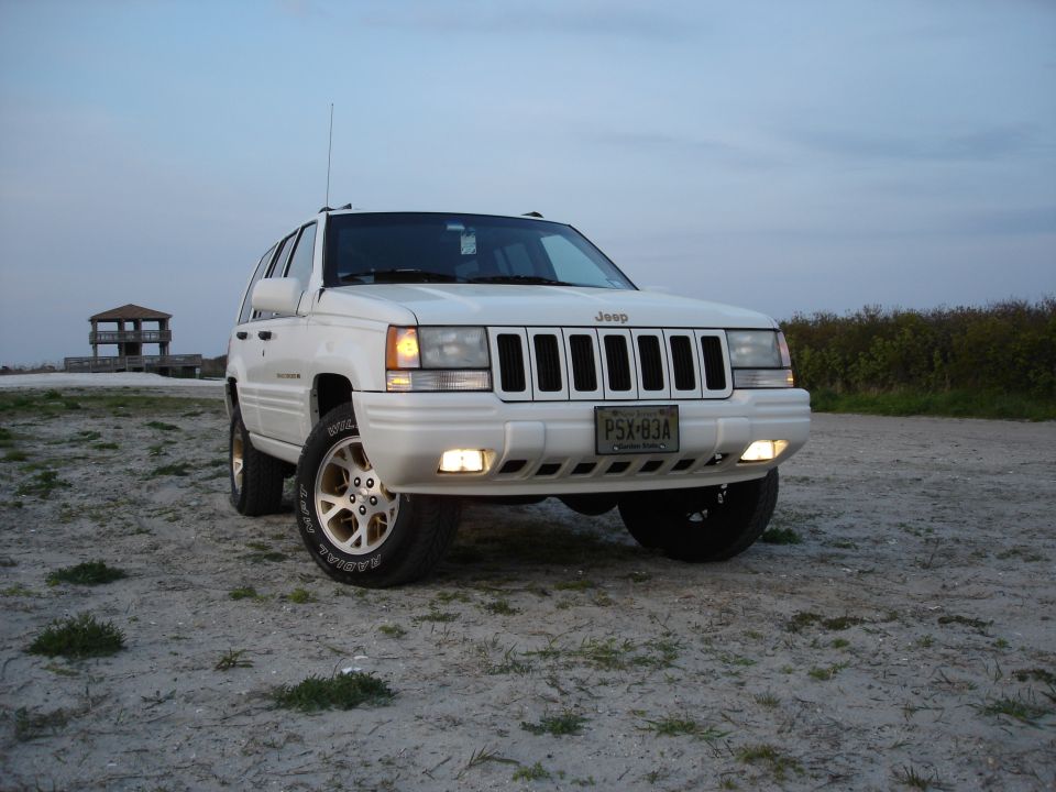 White Jeep Grand Cherokee ZJ with gold alloy wheels parked on sand at dusk, lights on
