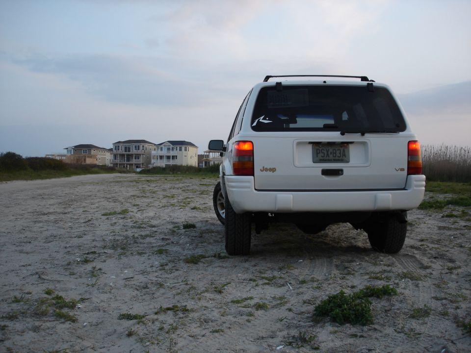 White Jeep Grand Cherokee rear view parked on sand near beach houses at dusk