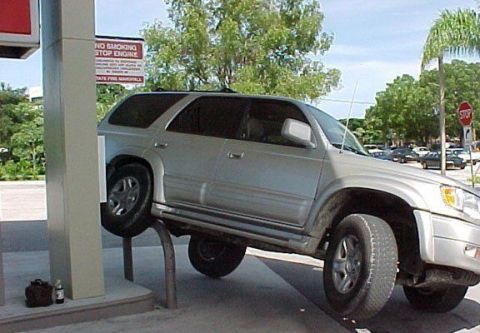 1985 Ford Bronco II - A silver SUV is precariously balanced on a gas pump bollard, with its front wheel elevated.