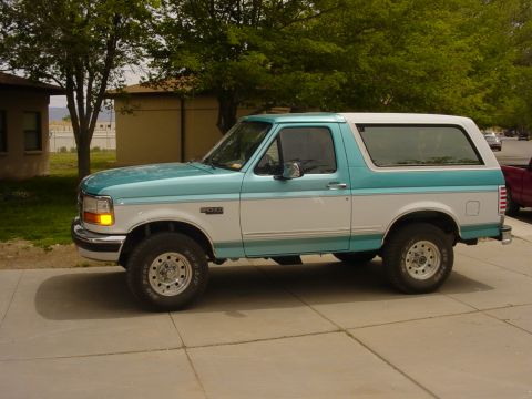 1995 Ford Bronco - A two-tone teal and white Ford Bronco is seen from the side, parked on a concrete driveway with trees and buildings in the background.