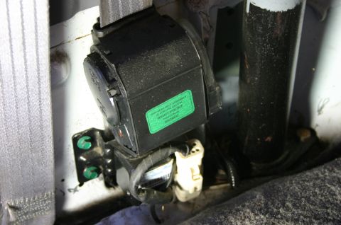1994 Ford Bronco - Close-up of a seat belt retractor assembly mounted at the pillar base with a green factory label reading "THIS SEAT BELT ASSEMBLY IS FOR US…