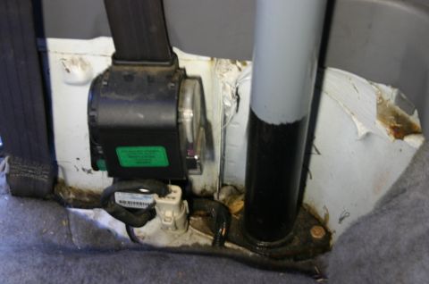 1994 Ford Bronco - Interior floor-level detail with the carpet pulled back, exposing a seat-belt retractor with a green warranty/inspection label, its white w…
