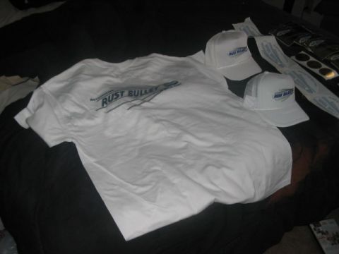 1991 Ford Bronco - A white t-shirt and two white trucker hats with the 'Rust Bullet' logo are laid out on a dark surface, alongside several stickers.