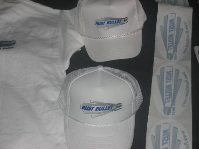 White t-shirt, two caps, and stickers with blue 'Rust Bullet' branding on a dark background.