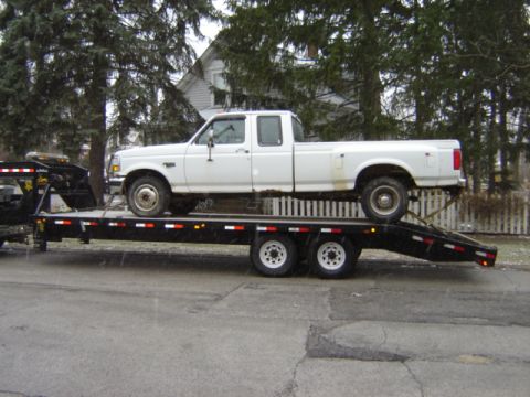 1992 Ford Bronco - A white pickup truck is loaded onto a flatbed trailer, parked on a street.