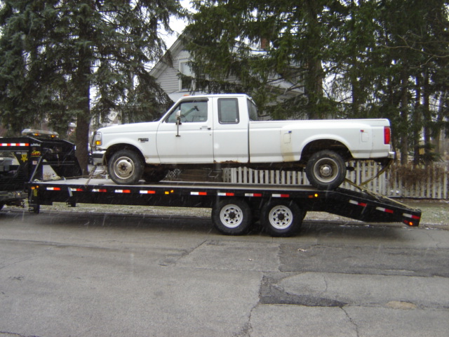 White crew cab pickup truck on a black trailer. Truck has four doors and a long bed