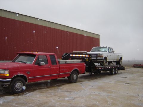 1992 Ford Bronco - Red Ford F-Series pickup tows a white Ford F-Series pickup on a trailer in front of a red building.