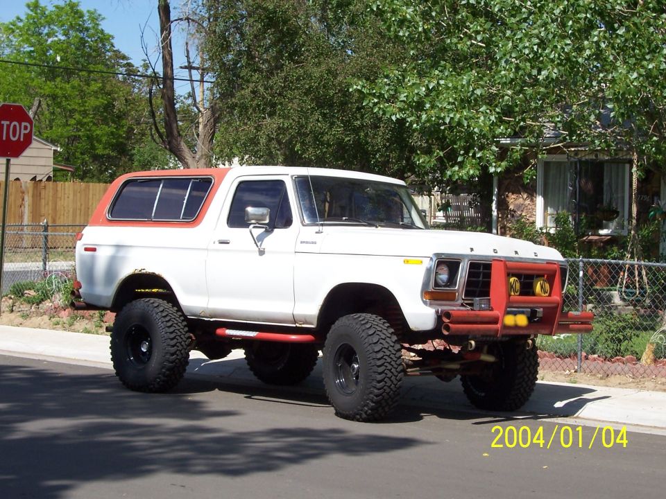 White 1978 Ford Bronco with orange roof and red custom bumper, lifted on large tires, parked on street