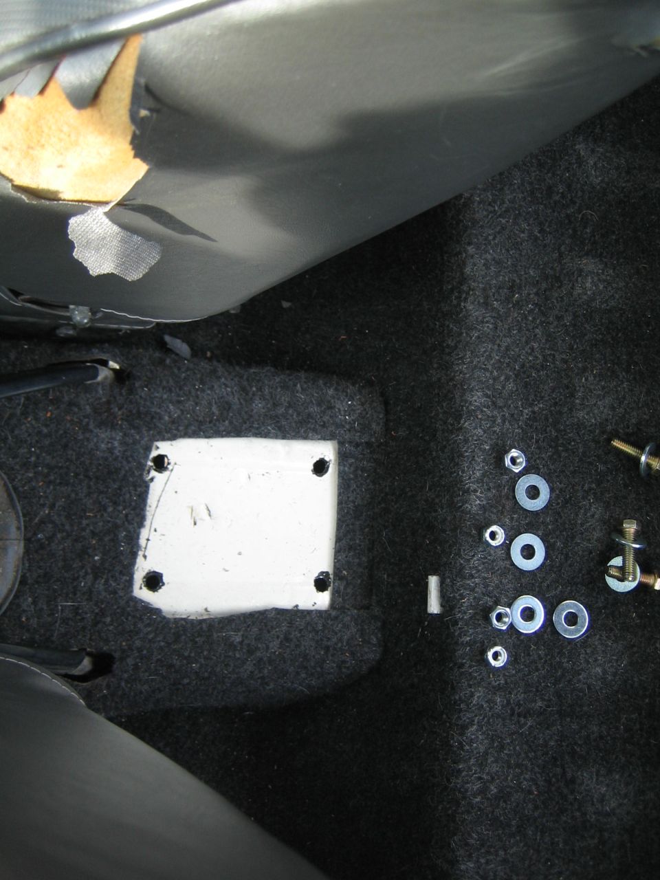 Torn gray seat upholstery above a white plate with four bolt holes on a dark carpeted floor next to scattered nuts and bolts