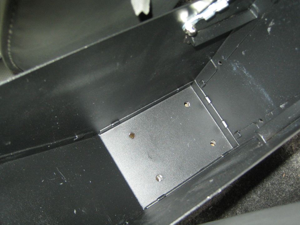Black metal storage box with four screw holes on the bottom plate, inside a vehicle