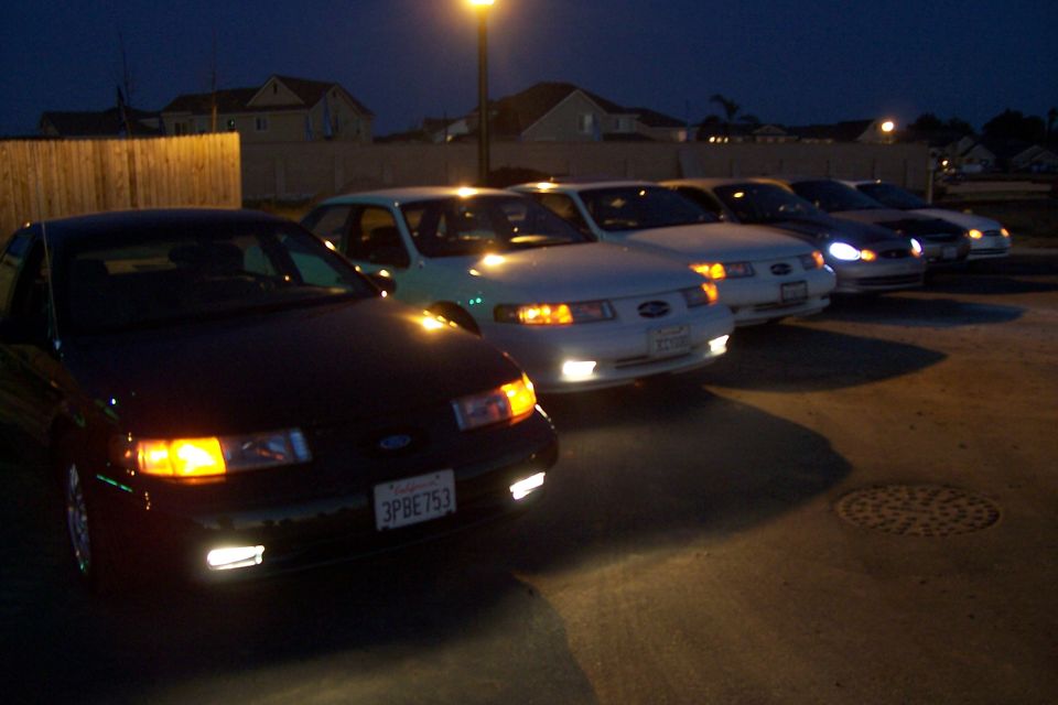 Several sedans parked in a line at night, headlights on. One car in foreground has license plate 3PBE753