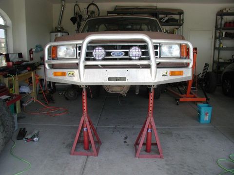 1991 Ford Bronco - The front of a Ford truck is elevated on jack stands in a garage, revealing its undercarriage and a large brush guard.