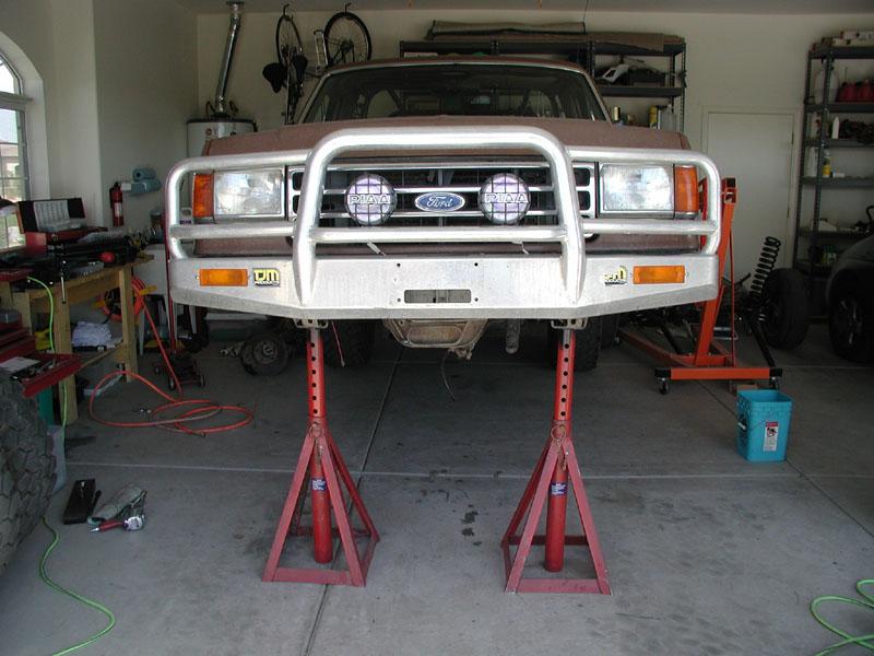Front of tan Ford truck on red jack stands with aluminum grille guard and PIAA lights in garage