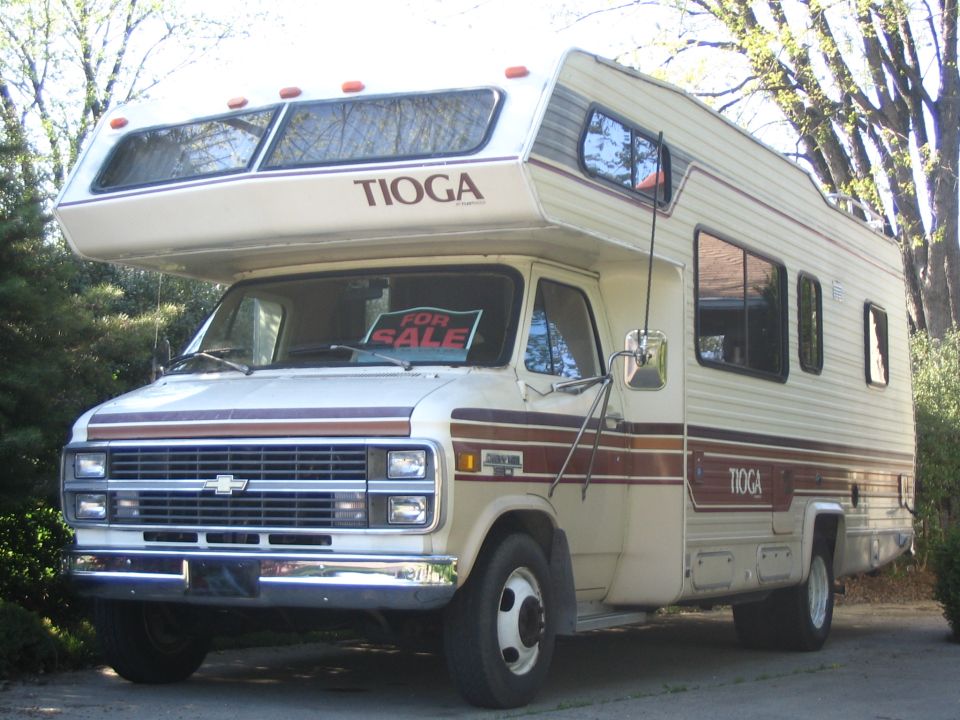 Cream Tioga Class C motorhome on Chevy chassis with For Sale sign in windshield, parked among trees