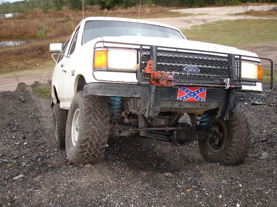 White Ford Bronco with a winch, brush guard, and Confederate flag on the front bumper. It has large off-road tires and blue suspension springs