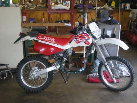 1900 Honda Big Red - A white and red Honda dirt bike is parked in a workshop. The 'CRF' logo is visible on the red seat.