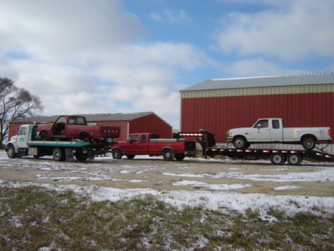 1992 Ford Bronco - Several trucks are being transported on tow trucks and trailers near red barns on a cloudy day.