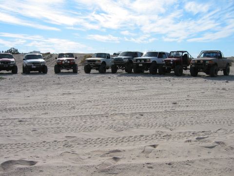 1997 Ford Ranger - A group of off-road trucks are parked on a sandy terrain under a cloudy sky, with one identified as a 1997 Ford Ranger.