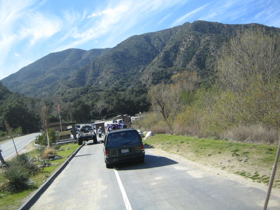 Several SUVs and off-road vehicles are parked near a mountain road with a sign and a flag