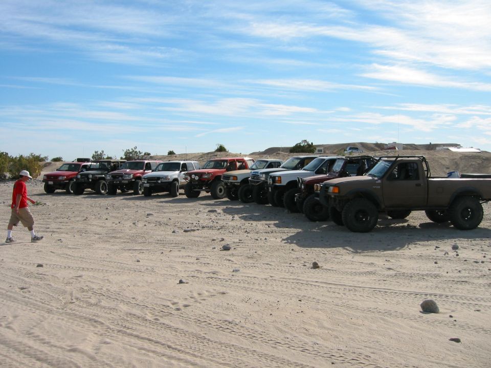 Several off-road trucks parked in a sandy lot with a person walking