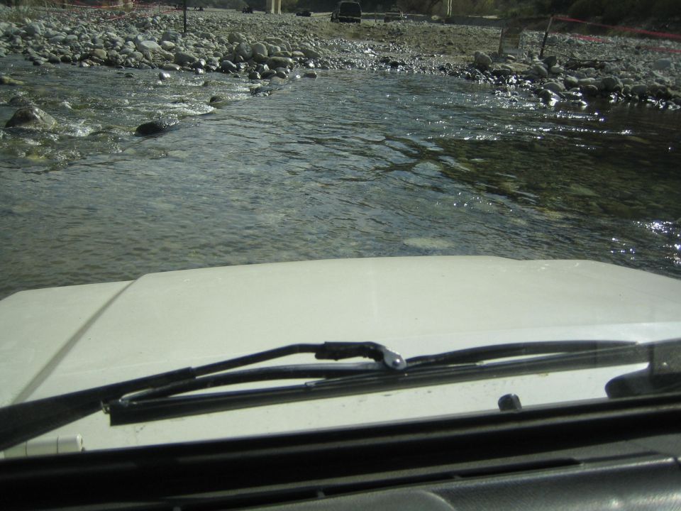 View from inside a vehicle crossing a rocky river, with another vehicle visible in the distance