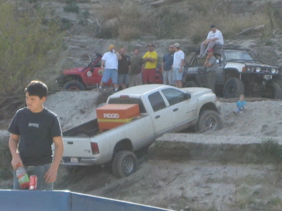 Several off-road vehicles are stuck in sand, with people watching. A white pickup truck is in the foreground