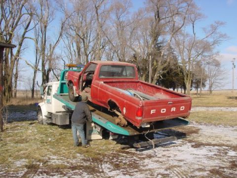 1992 Ford Bronco - A red Ford pickup truck is loaded onto a tow truck in a rural setting with bare trees and patchy snow.