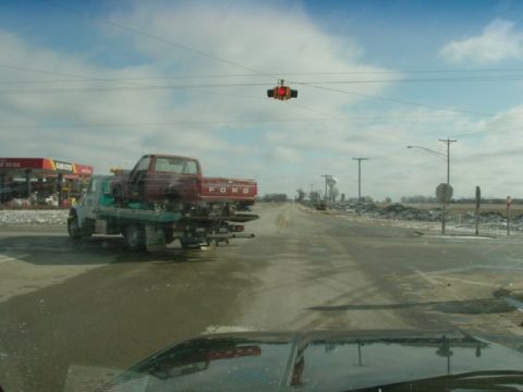 1992 Ford Bronco - A red Ford pickup truck is being towed on a flatbed truck past a gas station.