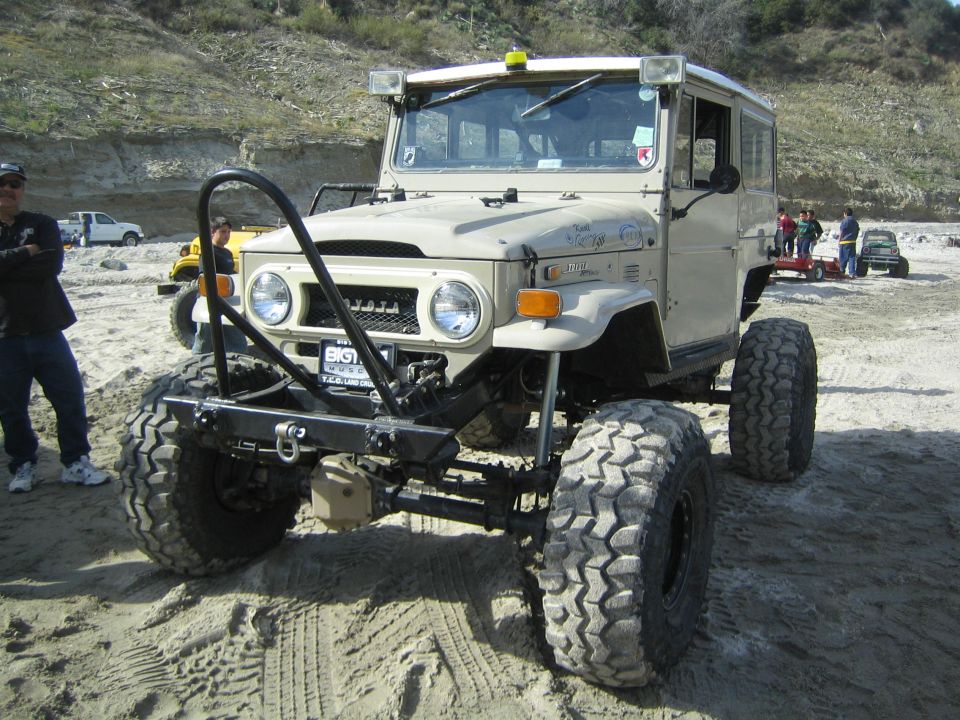 Tan Toyota Land Cruiser FJ40 with large off-road tires and a custom bumper. Other vehicles and people are in the background