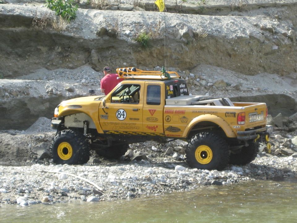 Yellow off-road pickup truck with large tires and roof rack, parked on rocky shore