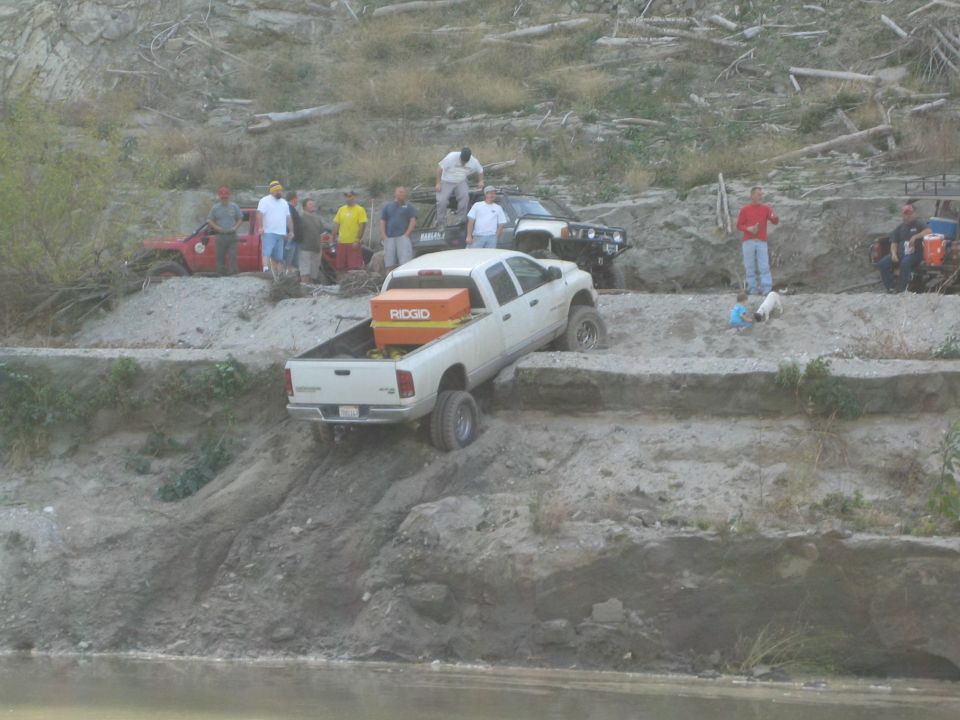 White pickup truck with orange Ridgid toolbox in bed, tilted on a dirt embankment next to a body of water. Other off-road vehicles and people are visible in the background